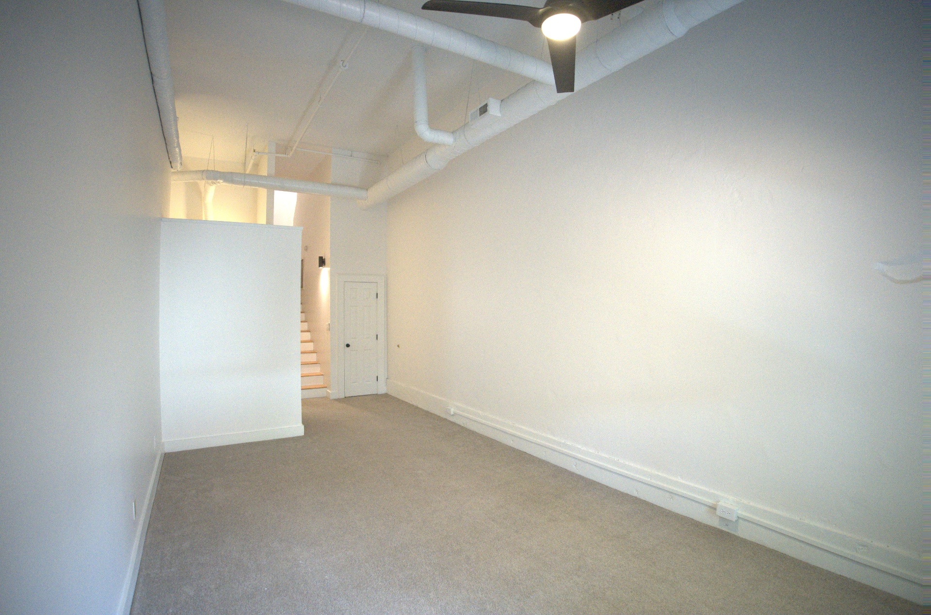 Empty loft bedroom with carpet, white walls, ceiling fan and exposed ceiling pipes