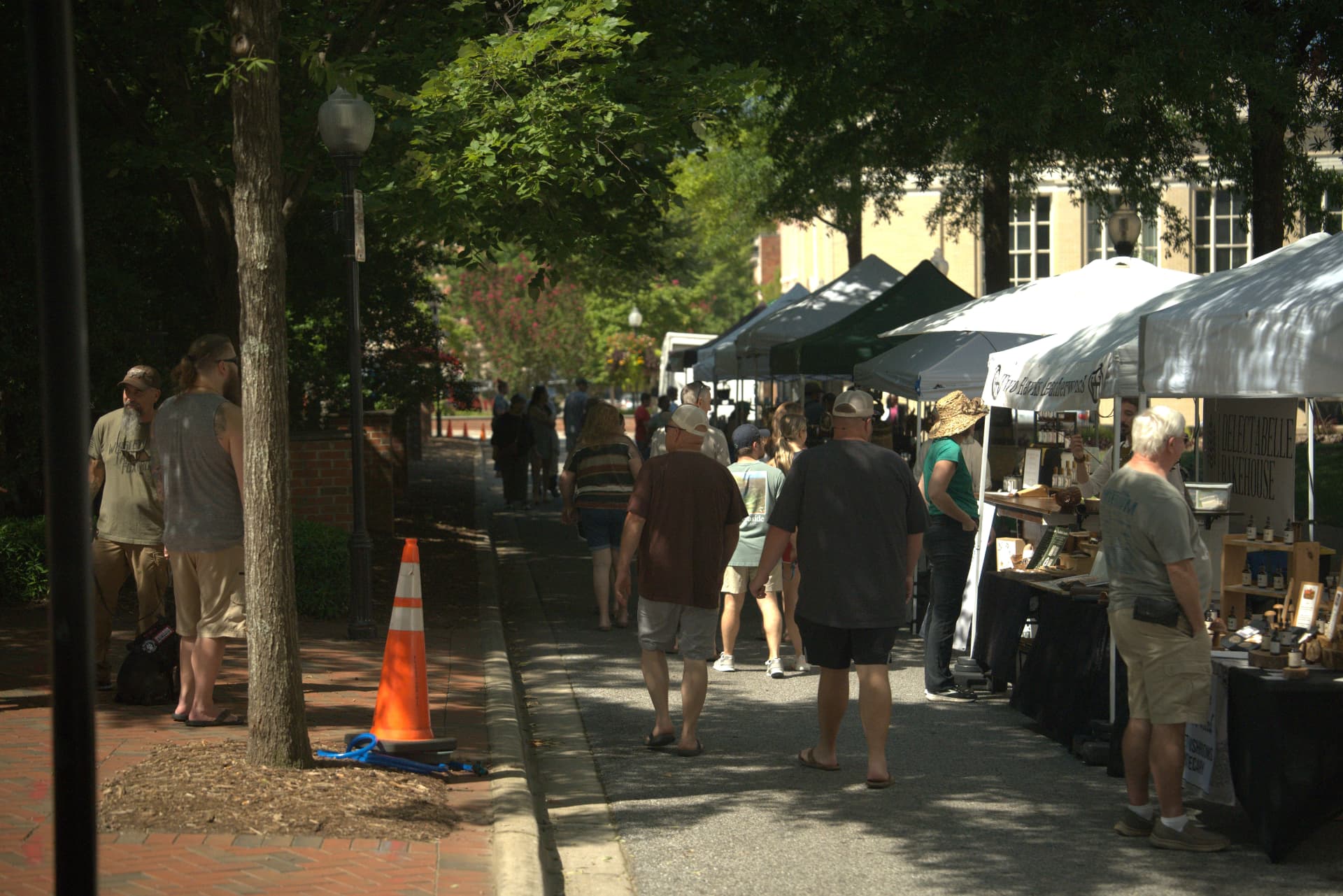 Shoppers walking a tree-shaded downtown Spartanburg sidewalk lined with white outdoor market tents and vendor tables